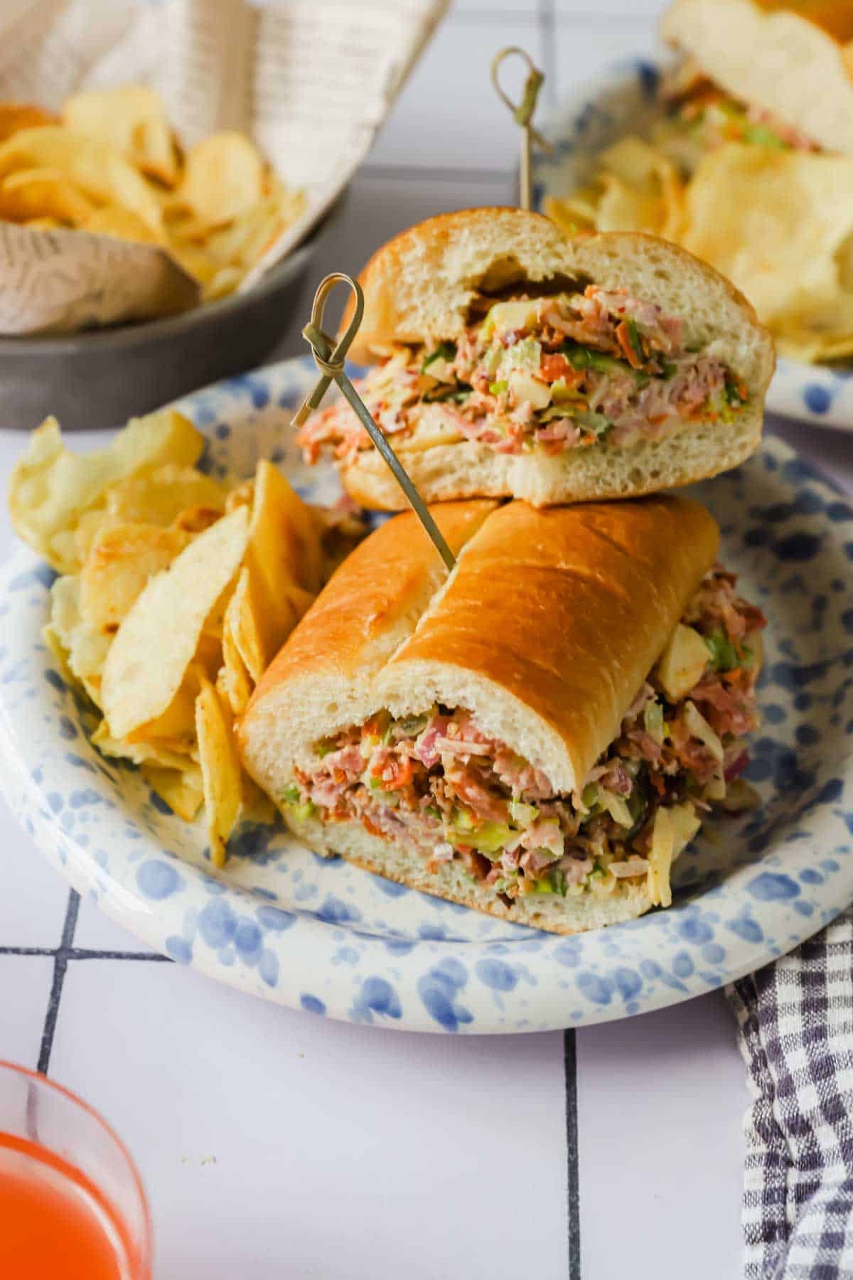 A sandwich filled with tuna salad and vegetables, sliced in half and held with skewers, served with potato chips on a patterned plate.