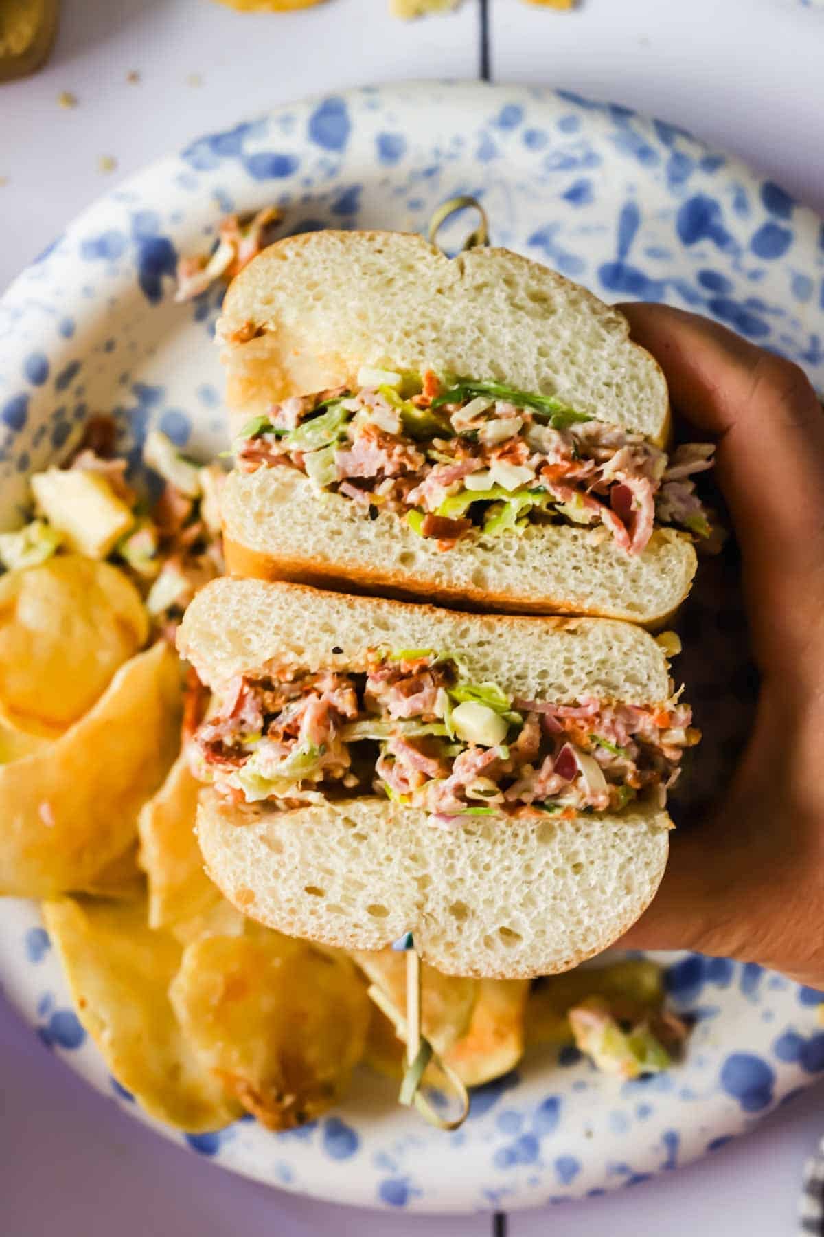 A hand holds a sandwich filled with shredded meat and lettuce, sliced in half on a plate with potato chips.