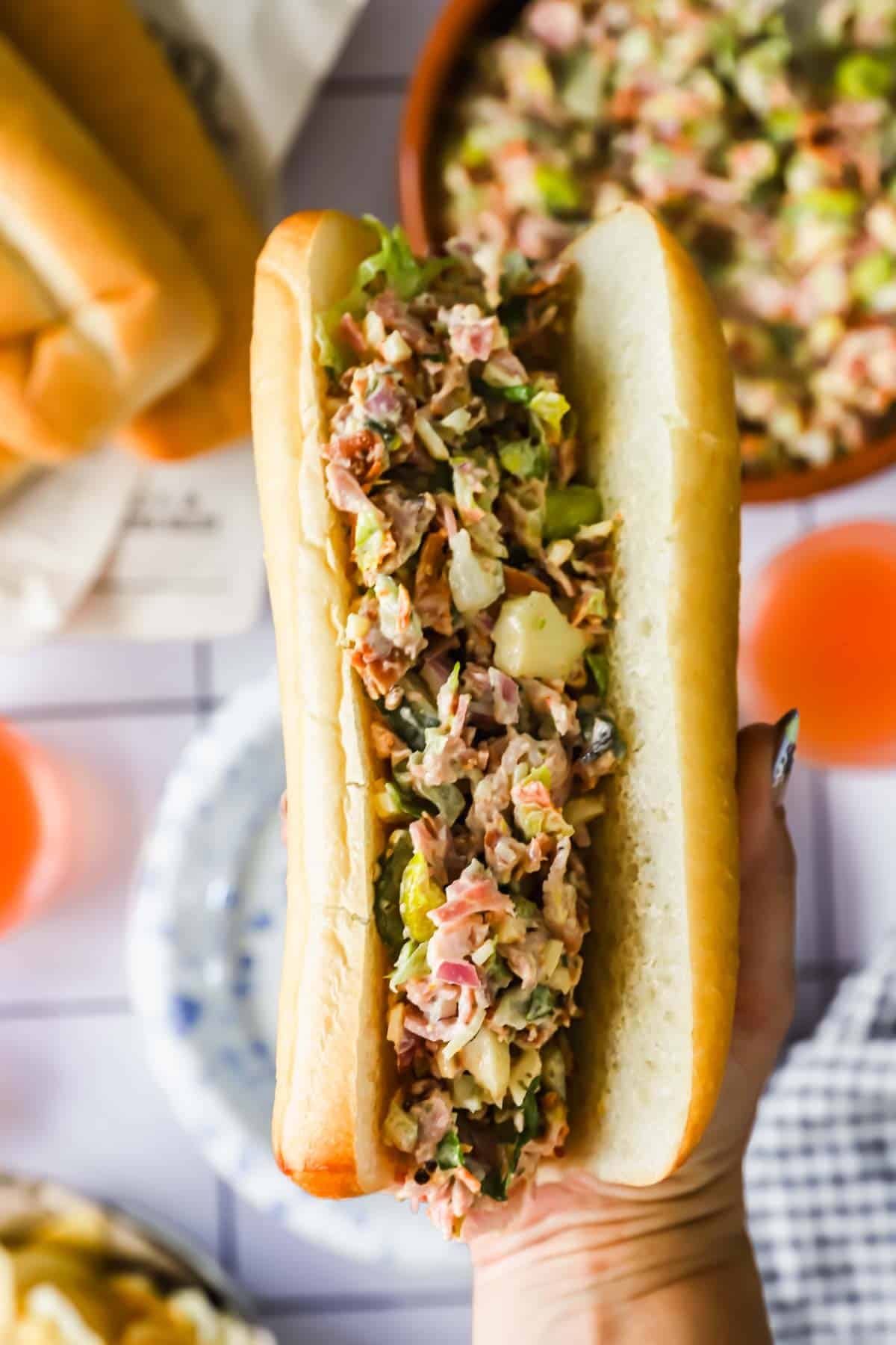 A hand holds a bun filled with chopped deli meat, cheese, lettuce, and pickles, with more filling and drinks visible in the background.