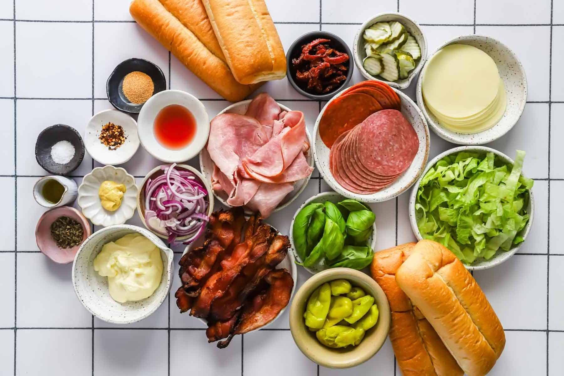 Various sandwich ingredients arranged on a white grid surface, including deli meats, cheeses, vegetables, sauces, seasonings, and sandwich rolls.
