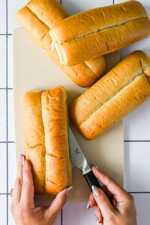 Four sandwich rolls on a cutting board; one is being sliced open with a knife by a person with manicured nails.
