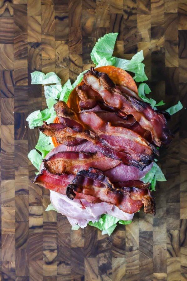 An open-faced sandwich on a wooden cutting board with layers of lettuce, tomato, sliced ham, and several strips of cooked bacon.