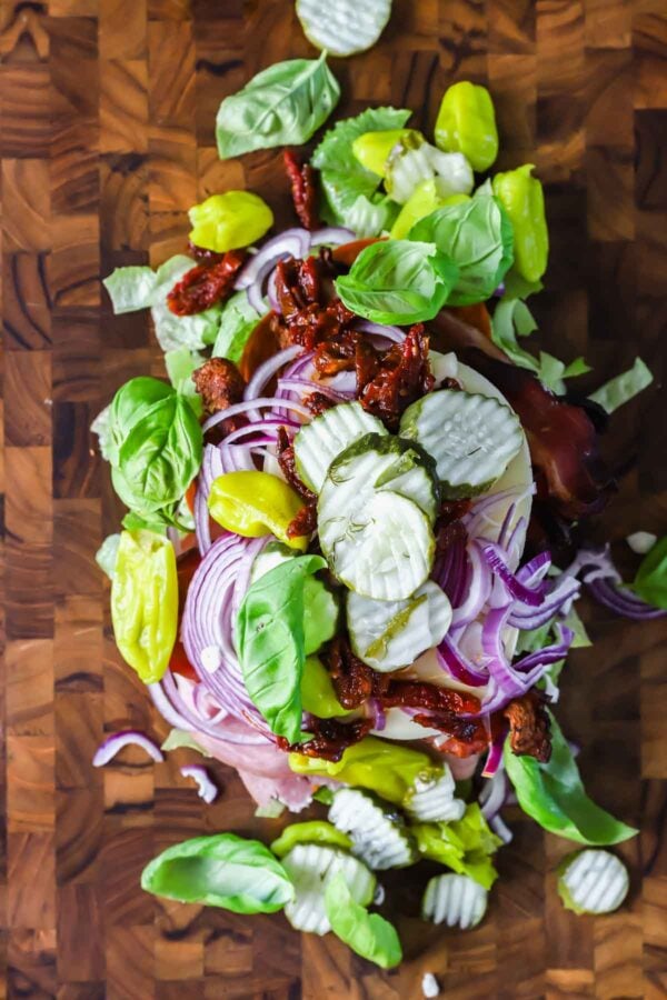 Sandwich ingredients including pickles, red onion slices, basil leaves, pepperoncini, and sun-dried tomatoes arranged on a wooden cutting board.