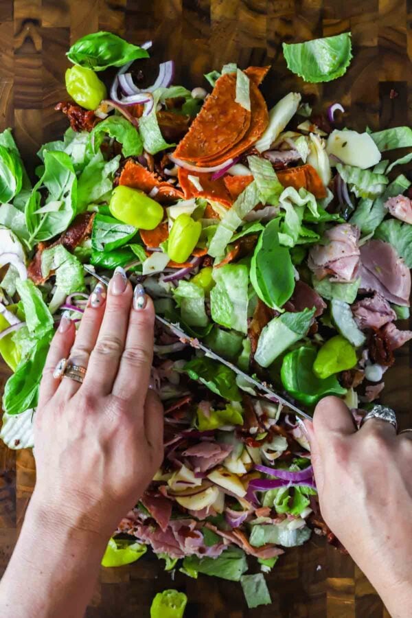 Two hands chop a pile of deli meats, lettuce, onions, pepperoncini, cheese, and pepperoni on a wooden cutting board.
