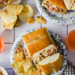Two plates with chopped sandwiches and potato chips, accompanied by drinks, sit on a white table. The image promotes "how to make the best chopped sandwich" from a food blog.