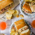 Three plates with sub sandwiches filled with mixed salad, potato chips on the side, and two glasses of orange drink on a white tiled surface.