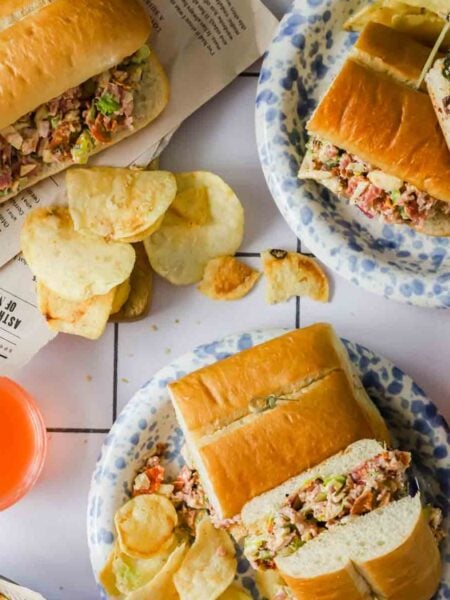 Three plates with sub sandwiches filled with mixed salad, potato chips on the side, and two glasses of orange drink on a white tiled surface.