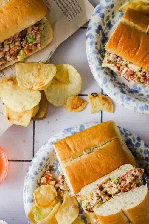 Three plates with sub sandwiches filled with mixed salad, potato chips on the side, and two glasses of orange drink on a white tiled surface.