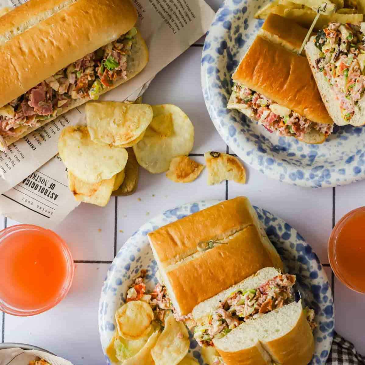 Three plates with sub sandwiches filled with mixed salad, potato chips on the side, and two glasses of orange drink on a white tiled surface.