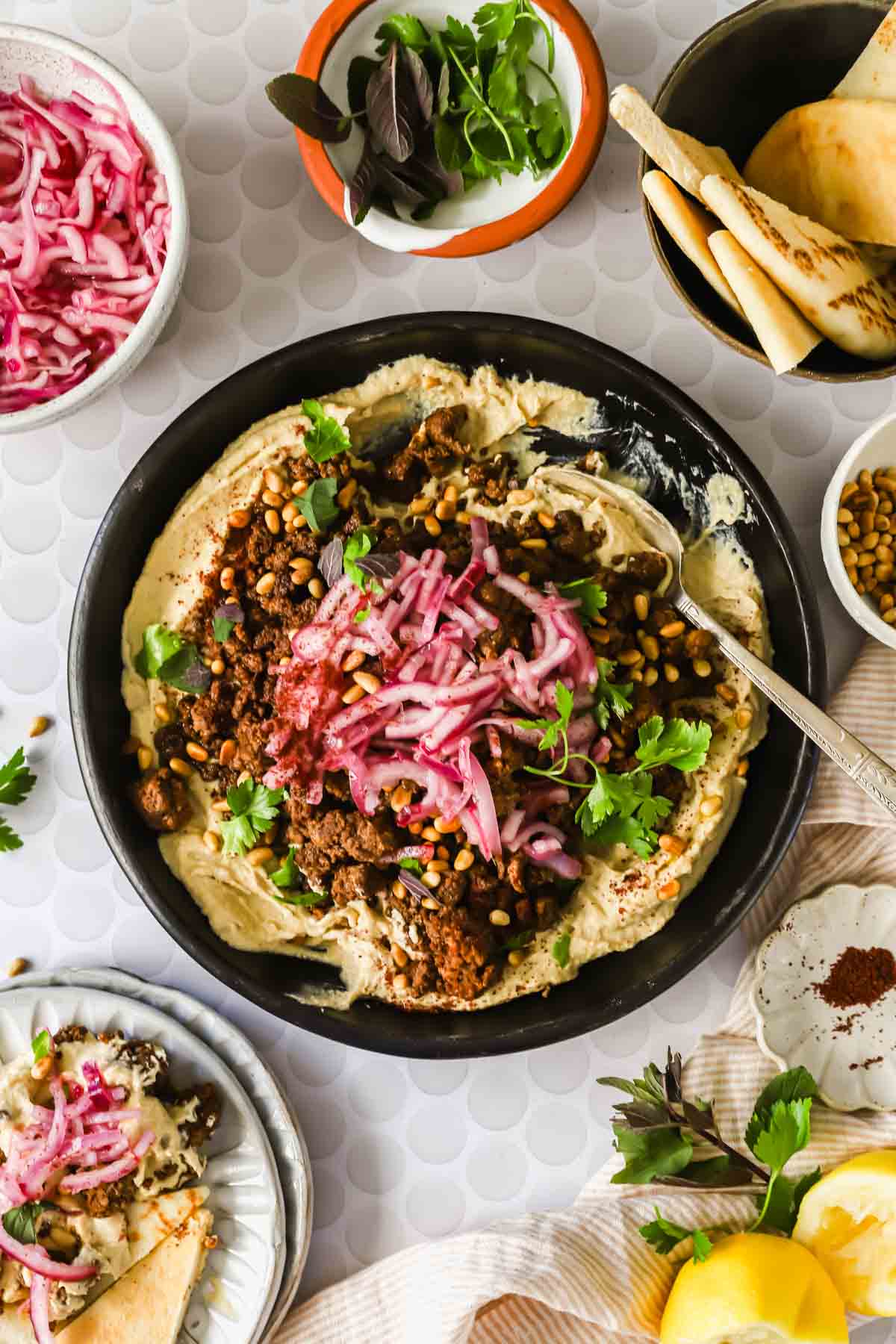 A bowl of hummus topped with ground meat, cooked lentils, pickled onions, pine nuts, and herbs, surrounded by bowls of pita bread, pickled onions, herbs, and lemon wedges.