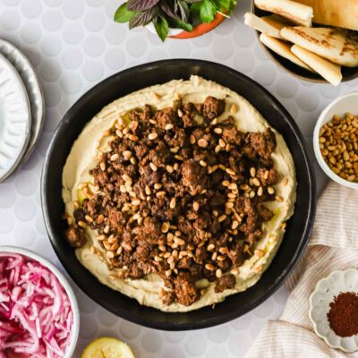 A bowl of hummus topped with spiced ground meat and pine nuts, surrounded by pita bread, herbs, lemon, pickled onions, and sumac on a light patterned surface.