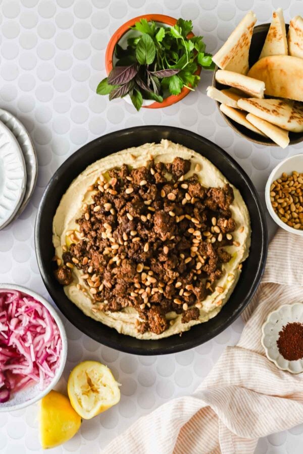 A bowl of hummus topped with spiced ground meat and pine nuts, surrounded by pita bread, herbs, lemon, pickled onions, and sumac on a light patterned surface.