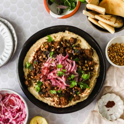 A bowl of hummus topped with ground meat, pine nuts, herbs, and pickled onions, surrounded by pita bread, lemon, fresh herbs, and small side dishes on a light patterned table.