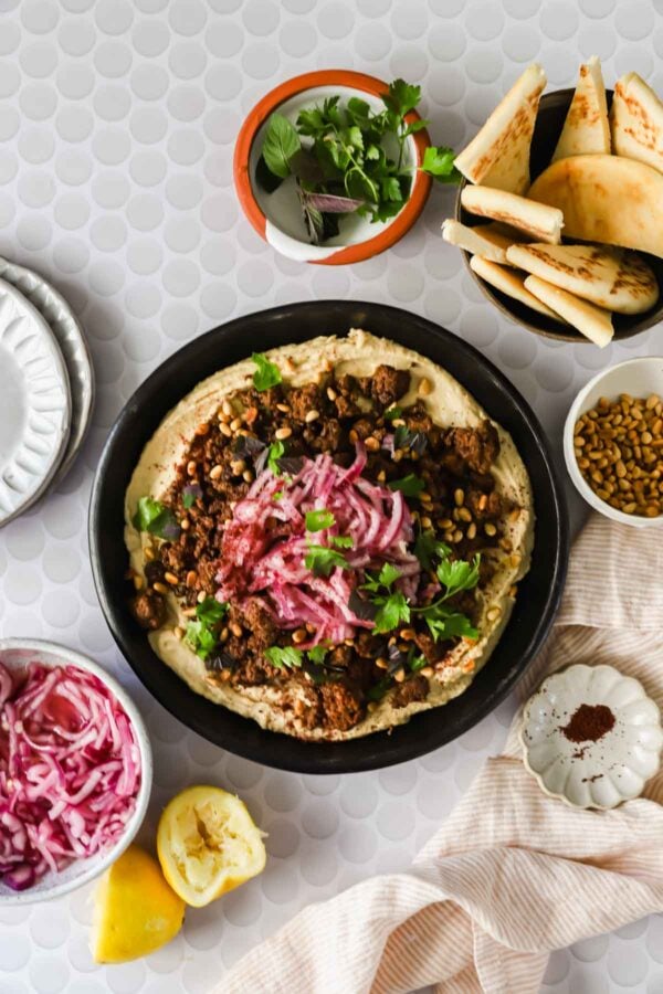 A bowl of hummus topped with ground meat, pine nuts, herbs, and pickled onions, surrounded by pita bread, lemon, fresh herbs, and small side dishes on a light patterned table.