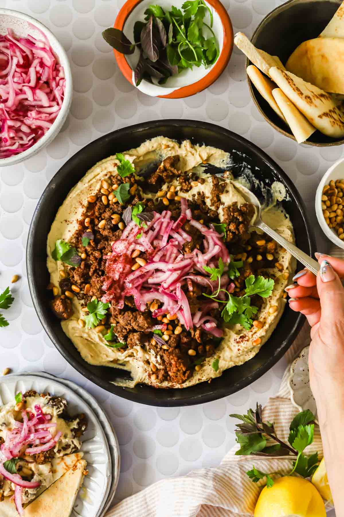 A bowl of hummus topped with seasoned ground meat, pine nuts, pickled onions, and fresh herbs, surrounded by pita, pickled onions, greens, and a hand holding a spoon.