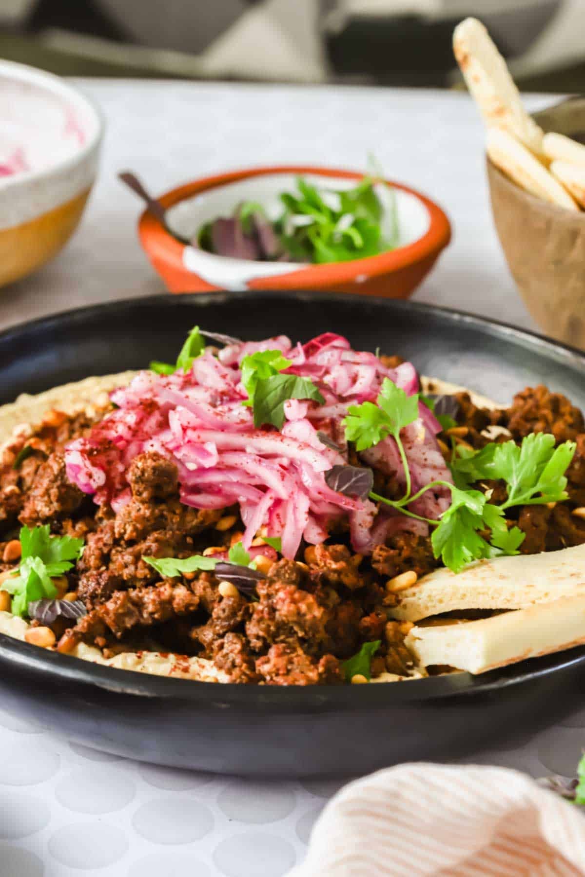 A black plate with seasoned ground meat, sliced onions, fresh parsley, and pita bread, with bowls of dip and extra bread in the background.