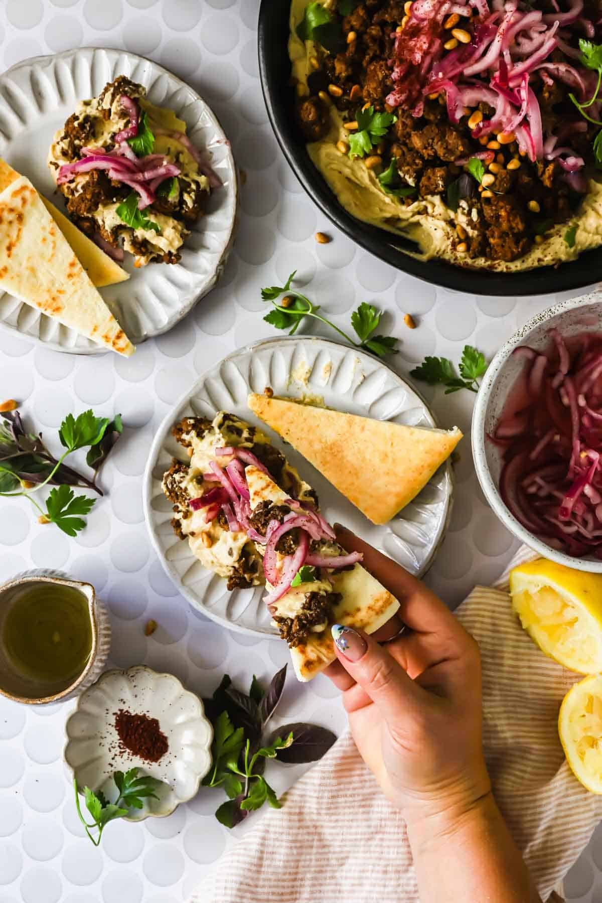 A hand holds a piece of flatbread topped with meat, pickled onions, and herbs. Plates with more flatbread, toppings, and lemon are on a white patterned table.