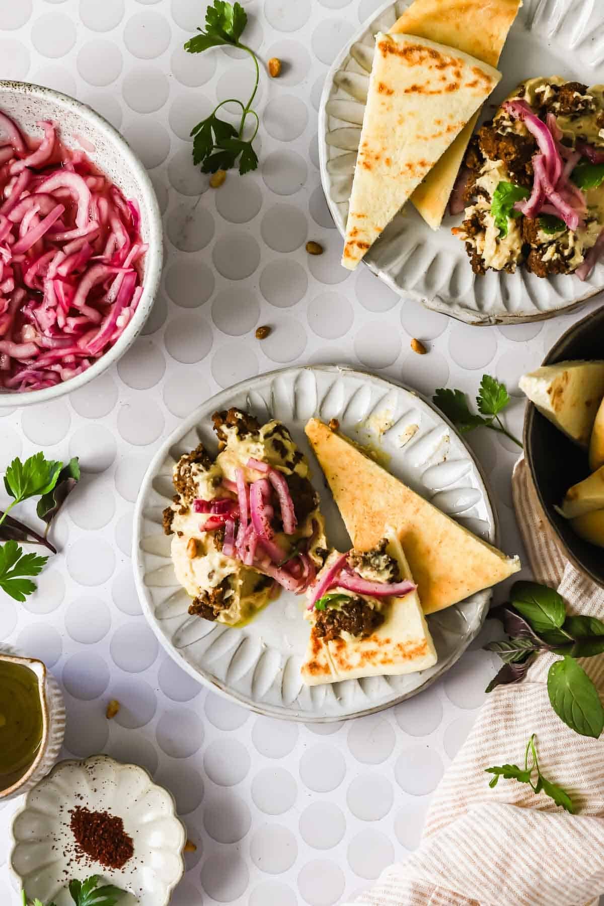 A plate with pita bread, hummus, and pickled red onions, surrounded by bowls of pickled onions, greens, and seasonings on a white textured surface.