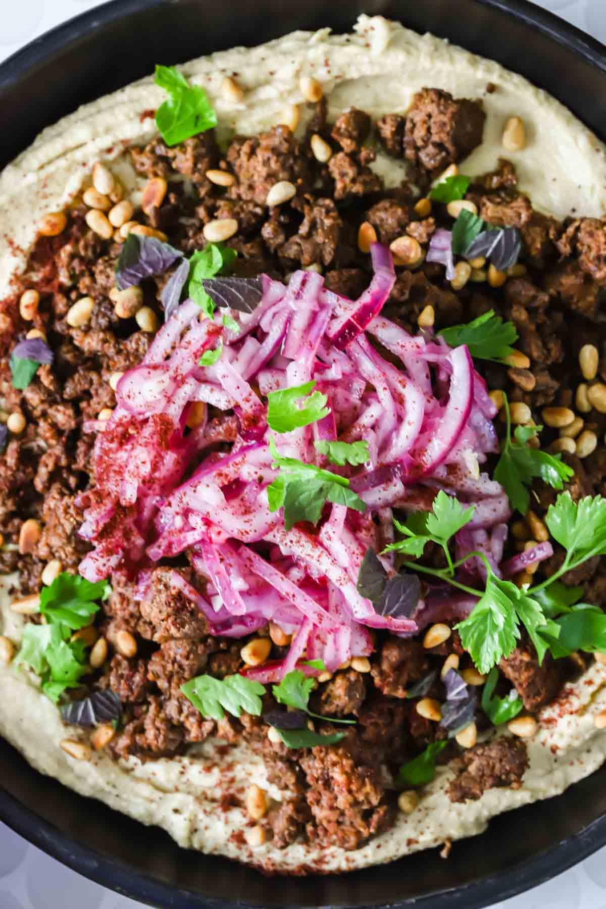 A black bowl filled with hummus, topped with ground meat, pickled red onions, pine nuts, fresh parsley, and spices.