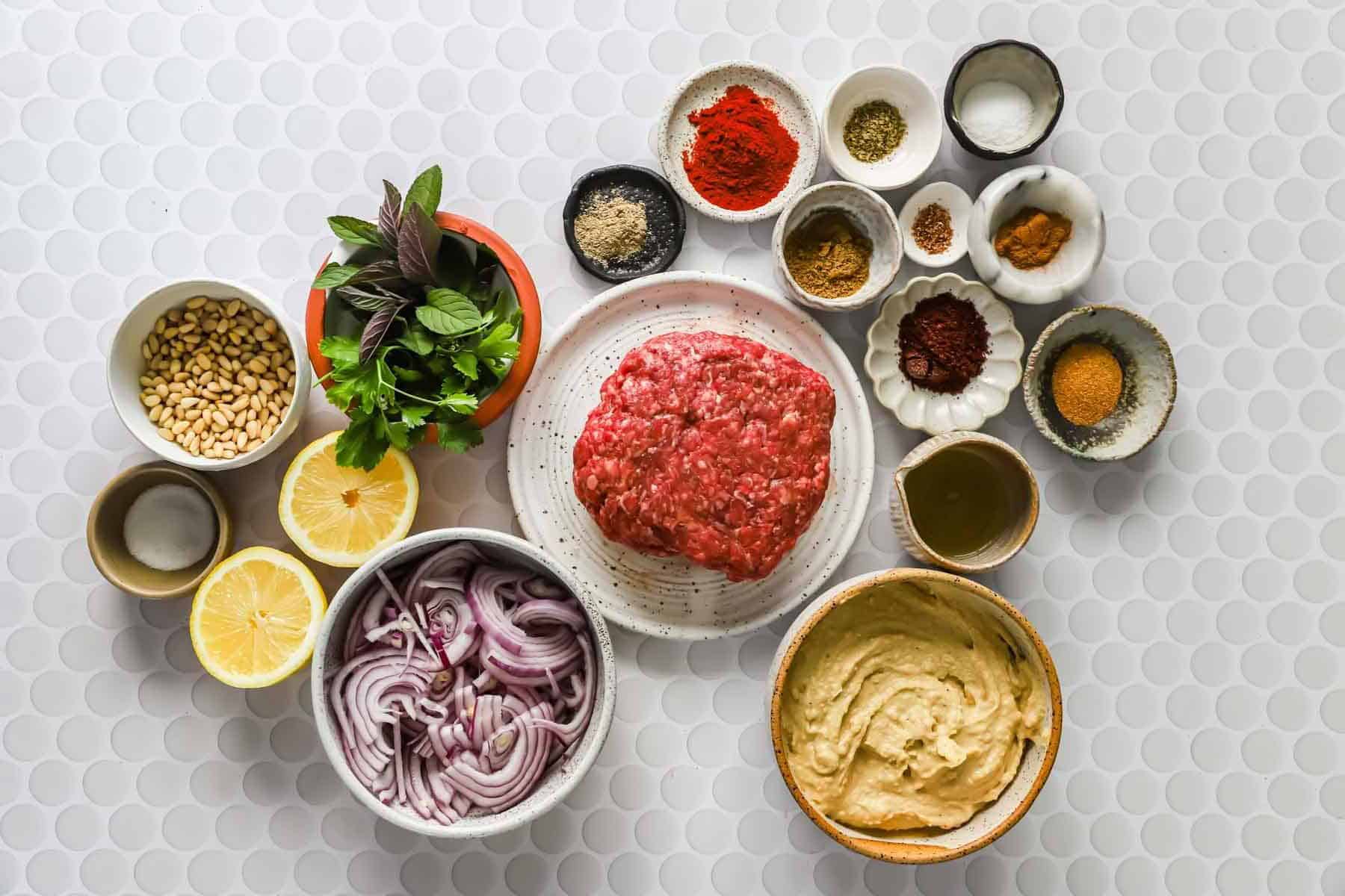 Various ingredients including ground meat, sliced red onions, hummus, spices, lemon halves, fresh herbs, pine nuts, and oil arranged on a white tiled surface.