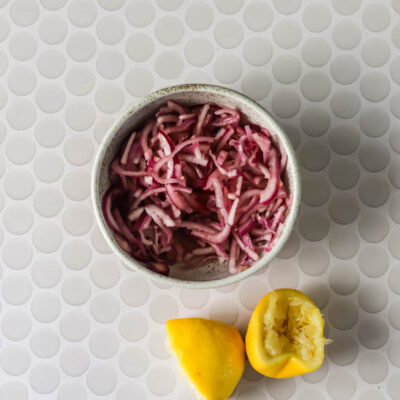 A bowl of shredded red onions sits next to two lemon halves, one of which is squeezed, on a white circular patterned surface.