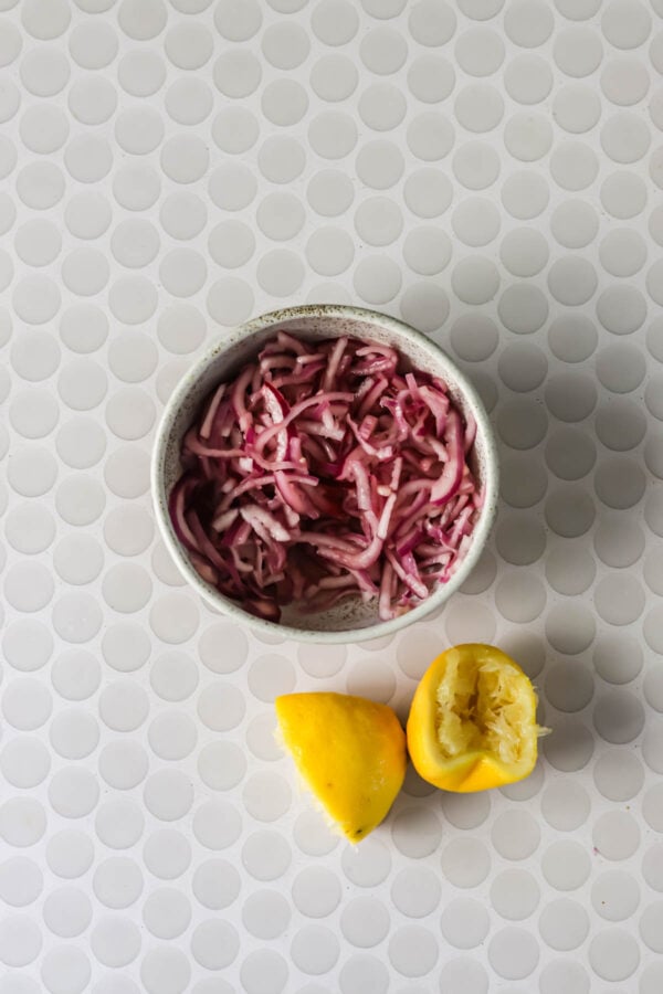 A bowl of shredded red onions sits next to two lemon halves, one of which is squeezed, on a white circular patterned surface.