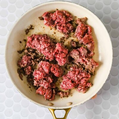 A white frying pan with partially browned ground beef sits on a white textured surface.