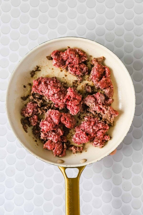 A white frying pan with partially browned ground beef sits on a white textured surface.