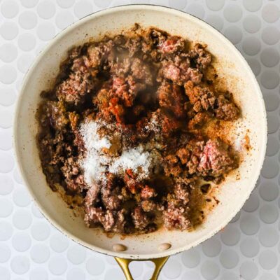 Ground beef cooking in a skillet, topped with various seasonings including salt and paprika, on a white patterned surface.