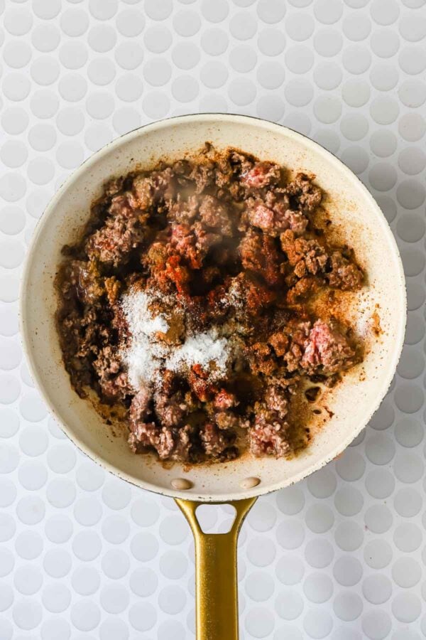 Ground beef cooking in a skillet, topped with various seasonings including salt and paprika, on a white patterned surface.