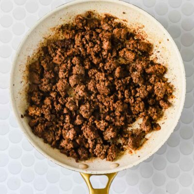Cooked ground beef in a white skillet with a gold handle, set on a white patterned surface.