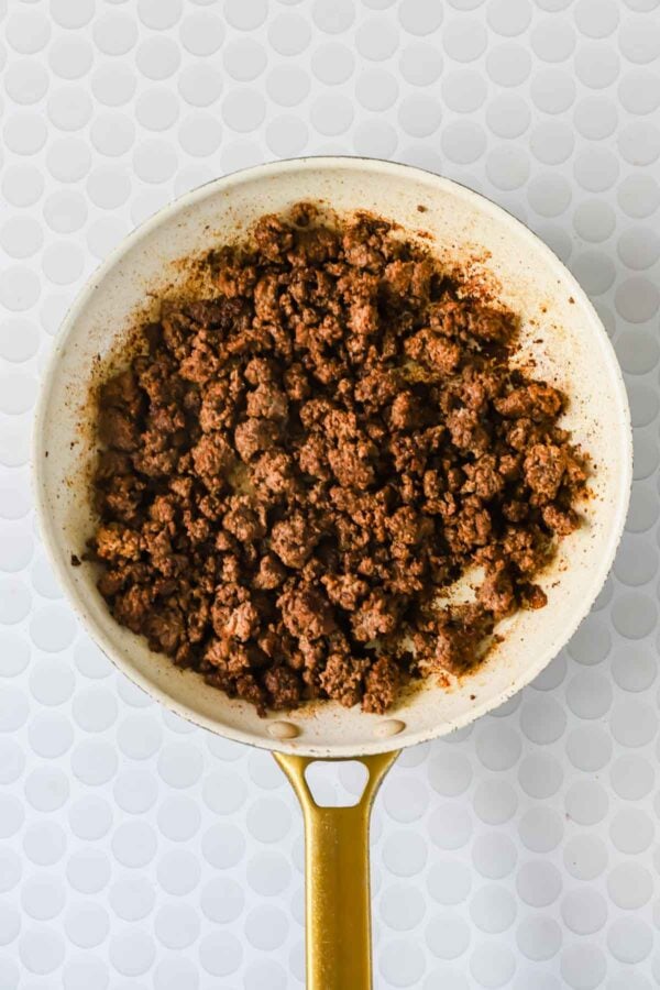 Cooked ground beef in a white skillet with a gold handle, set on a white patterned surface.