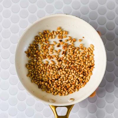 A white frying pan with golden brown toasted pine nuts, placed on a white surface with a circular pattern.