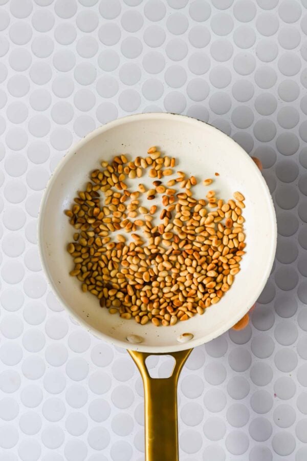 A white frying pan with golden brown toasted pine nuts, placed on a white surface with a circular pattern.