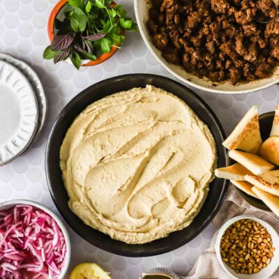 Ingredients for a Middle Eastern dish arranged on a table: a bowl of hummus, cooked ground meat, herbs, pita bread, sliced red onion, pine nuts, lemon, olive oil, and spices.