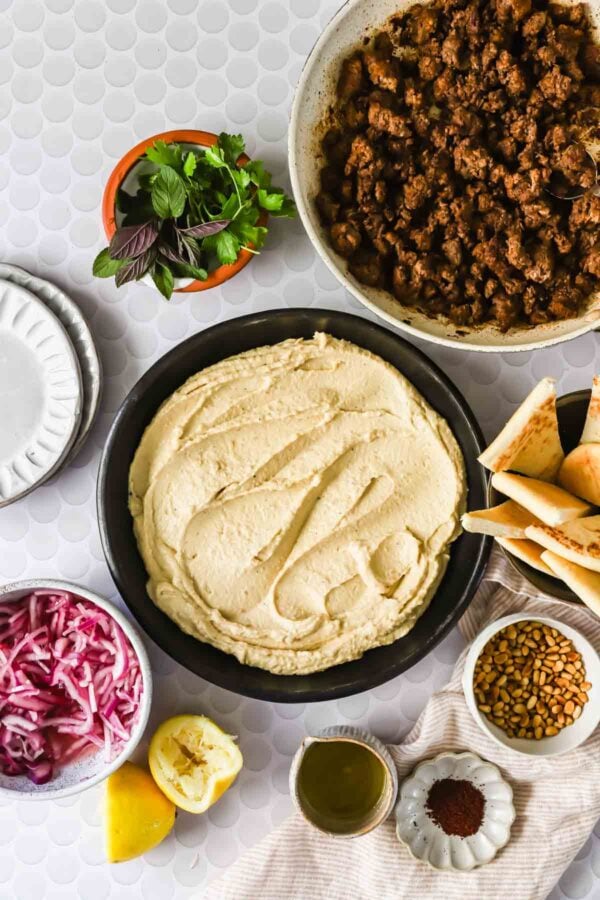 Ingredients for a Middle Eastern dish arranged on a table: a bowl of hummus, cooked ground meat, herbs, pita bread, sliced red onion, pine nuts, lemon, olive oil, and spices.