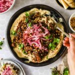 A bowl of hummus topped with spiced ground meat, pickled onions, pine nuts, fresh herbs, and a hand holding a spoon, with side dishes visible around the bowl.