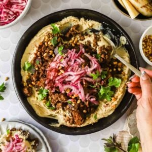 A bowl of hummus topped with spiced ground meat, pickled onions, pine nuts, fresh herbs, and a hand holding a spoon, with side dishes visible around the bowl.