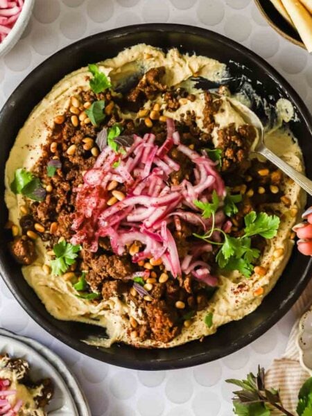 A bowl of hummus topped with spiced ground meat, pickled onions, pine nuts, fresh herbs, and a hand holding a spoon, with side dishes visible around the bowl.