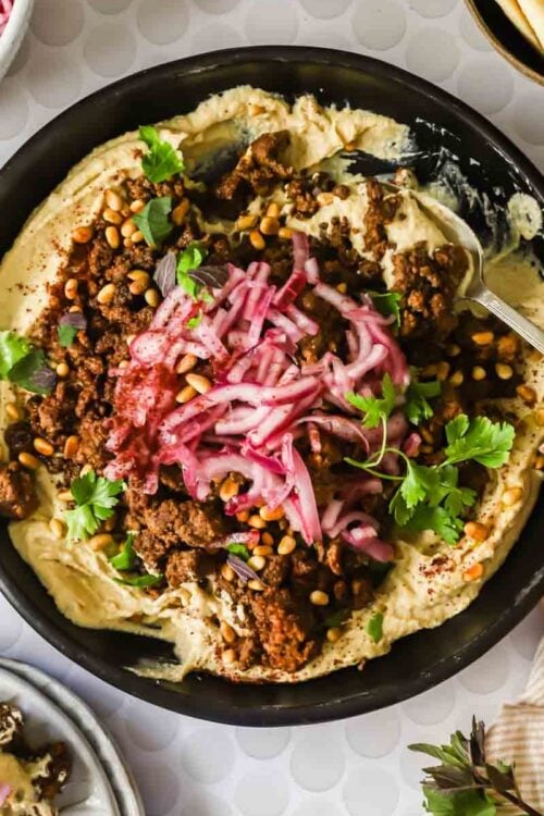 A bowl of hummus topped with spiced ground meat, pickled onions, pine nuts, fresh herbs, and a hand holding a spoon, with side dishes visible around the bowl.