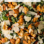A bowl of kale Caesar salad topped with croutons, shaved Parmesan cheese, and bacon, with text overlay reading "Kale Caesar Salad.