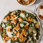 A bowl of kale Caesar salad topped with croutons and shaved parmesan, served with two forks.