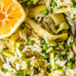 Close-up of orzo pasta salad with green peas, artichoke hearts, chopped basil, and a halved lemon, with a spoon visible in the dish.