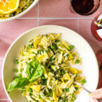 A bowl of lemon basil pasta salad with peas and artichokes sits on a pink tile surface, next to a drink and a larger bowl of salad.