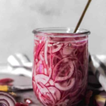 A glass jar filled with sliced pickled red onions sits on a counter with onion peels and a spoon inside. Text above reads "Best Pickled Onions.