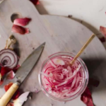 A glass jar of pickled red onions with a fork sits on a cutting board next to a sliced red onion and a knife.