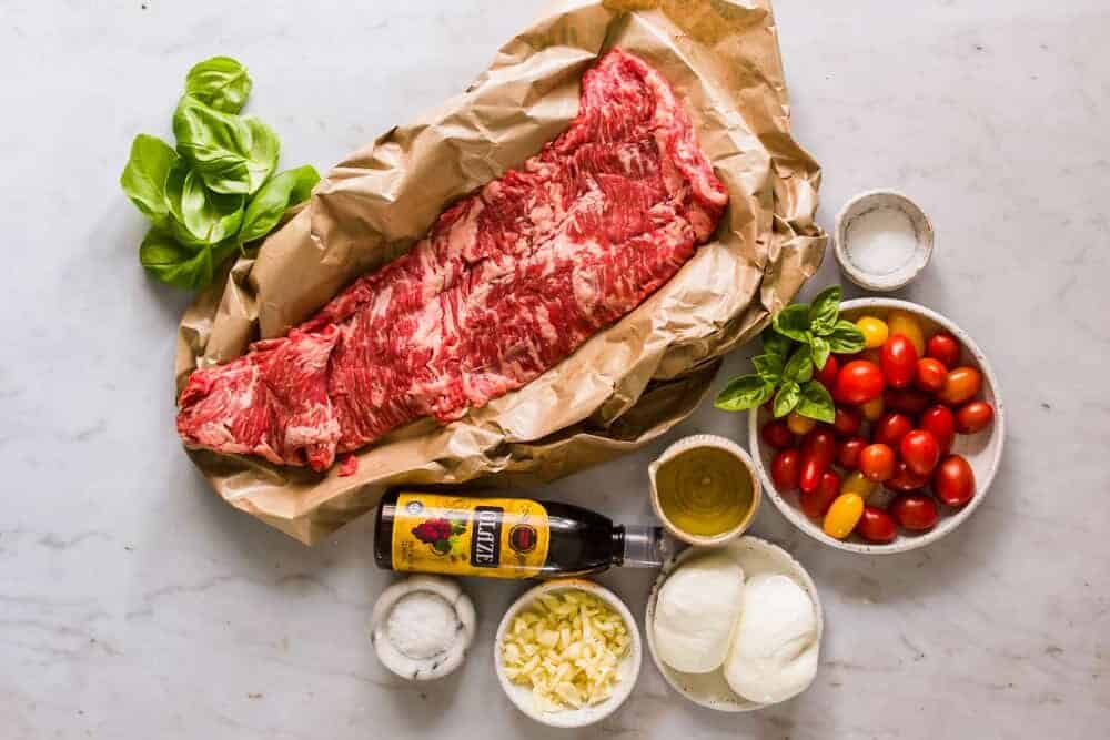 raw cut of skirt steak in brown paper next to white bowls with tomatoes, cheese, oil, garlic, and salt.