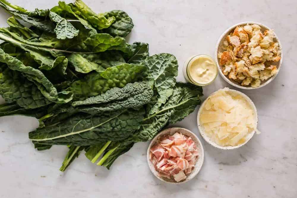 torn bread pieces, shaved cheese, and raw bacon in small white bowls next to kale