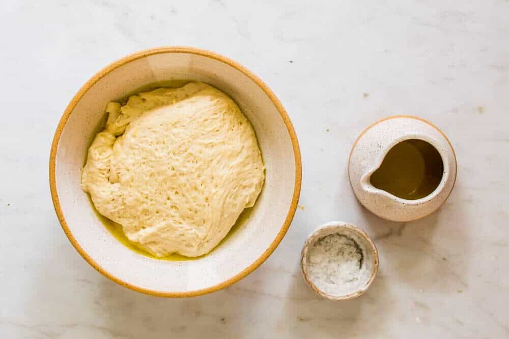 raw bread dough in a white bowl next to oil and salt