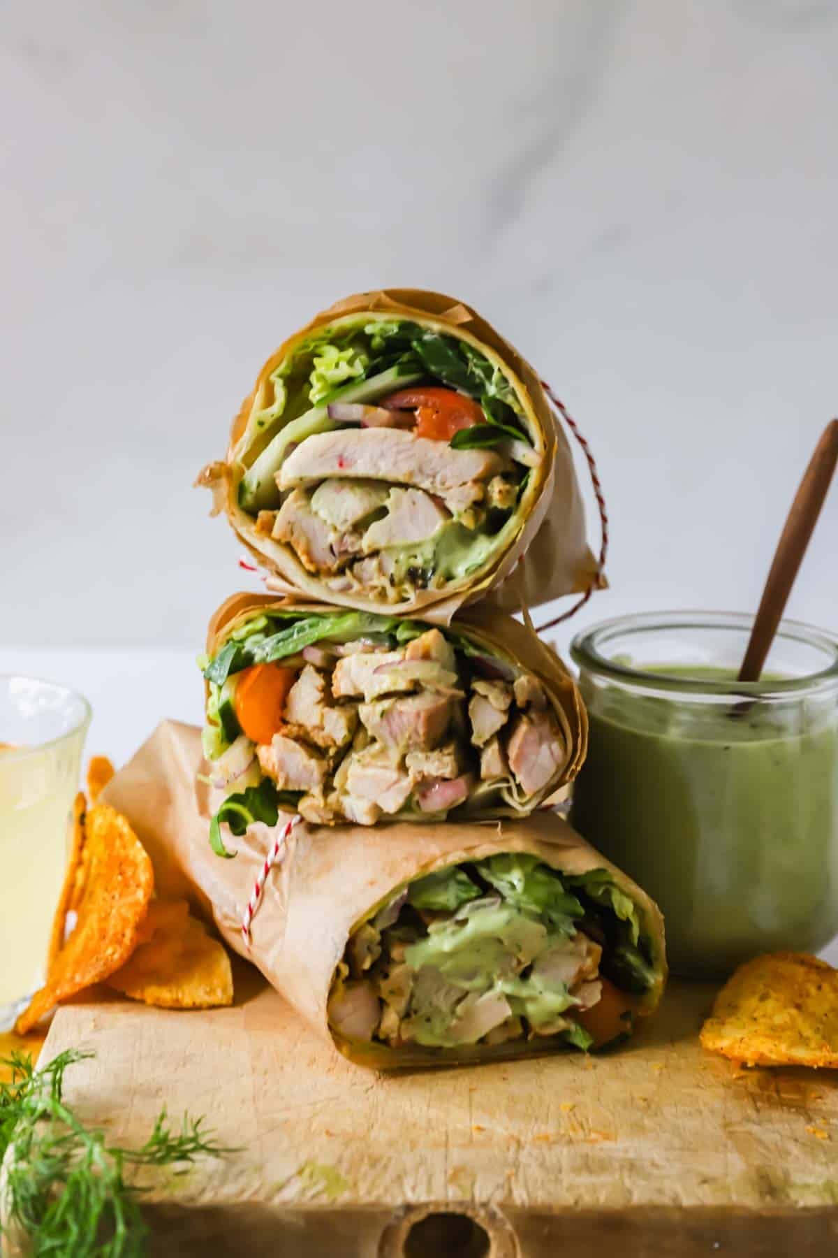 Three grilled chicken wraps filled with lettuce and tomato are stacked on a wooden board next to a glass of dipping sauce, a drink, and a few potato chips.
